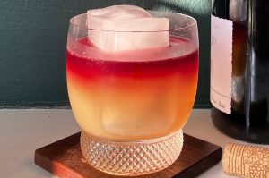 A cocktail fading from red to yellow in a glass next to a wine bottle
