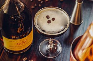 An Espresso martini cocktail on a dark bar top with a bottle of Cognac