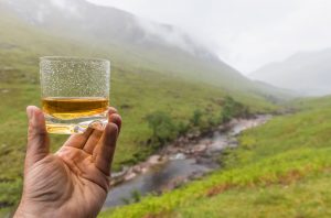 Hand holding glass of whisky with Scottish landscape in background